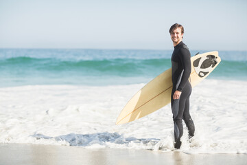 Adult male surfer wearing wetsuit with surfboard standing at shoreline as waves lap, copy space