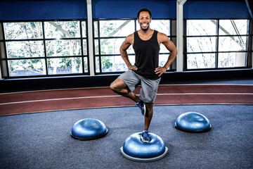 Man is balancing on one leg atop blue BOSU trainer in gym, with curved running track