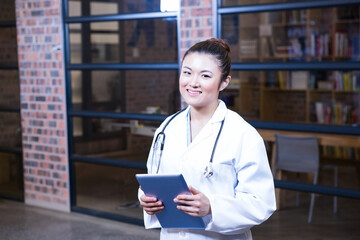 Asian female doctor standing in office holding tablet and wearing stethoscope by wooden bookshelves