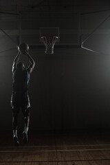 Basketball flying toward hoop in dimly lit gymnasium, spotlights casting dramatic shadows
