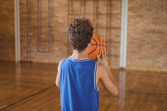 Boy dribbling basketball across polished wooden court inside school gym by brick wall with pipes