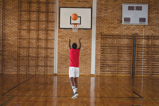 Asian teenage boy shooting basketball toward backboard-mounted hoop at school gym with scoreboard