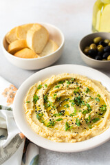 A bowl of creamy hummus topped with olive oil and herbs sits beside fresh bread and a bowl of olives. The light setting showcases a simple, inviting meal