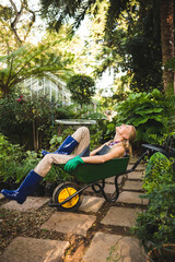 Green wheelbarrow is resting on stone pathway amid lush foliage with greenhouse peeking through