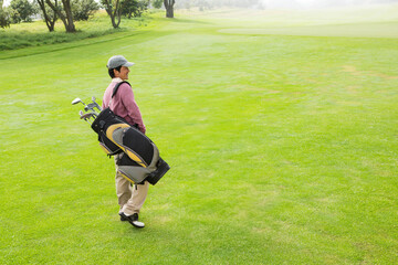 Obraz premium Mid adult Japanese man in golf attire walking across fairway carrying bag with clubs, copy space