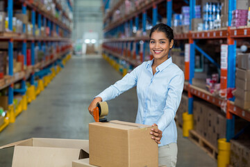 Packing station sealing cardboard box using tape dispenser in warehouse aisle with metal racks