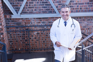 doctor wearing stethoscope climbing metal stairs beside red brick wall reviewing documents in sun