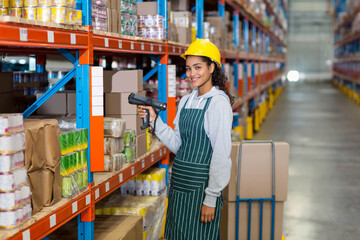 Female warehouse worker using handheld scanner scanning boxes on orange-blue racks in aisle by cart