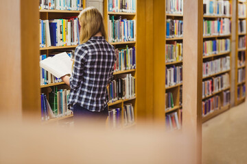 Female teenager standing in library aisle reading open hardcover book between wooden bookshelves