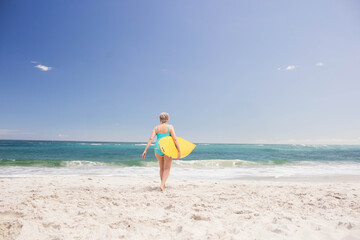 Senior woman walking into turquoise ocean on beach holding yellow surfboard in turquoise swimsuit