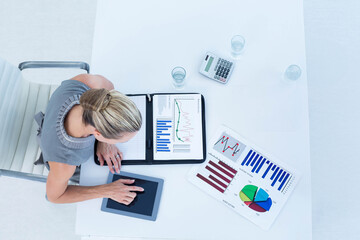 Middle-aged female wearing gray blouse studying financial charts on desk at office with tablet
