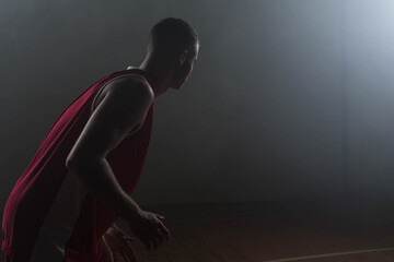 Basketball player is crouching on court under hazy spotlight, red jersey and floor markings shining