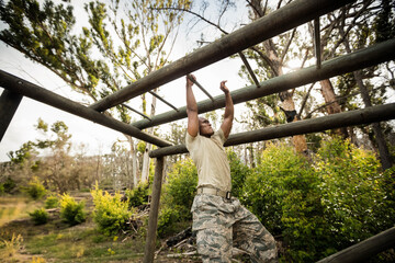 Male soldier wearing camouflage uniform gripping monkey bars and pulling on wooded training course