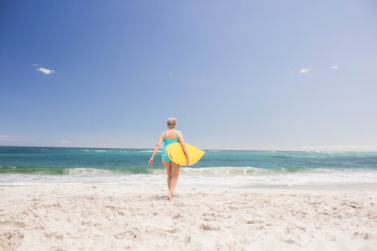 Senior woman walking into turquoise ocean on beach holding yellow surfboard in turquoise swimsuit