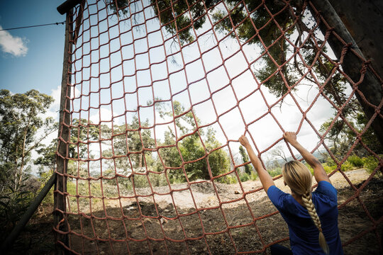 Woman gripping red rope net on wooden posts, preparing to climb wooded obstacle course, copy space