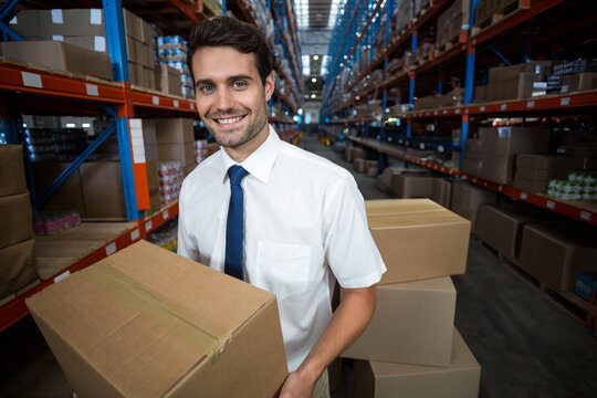Warehouse worker holding medium brown box in aisle with blue shelving, stacked boxes behind