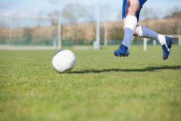 Teen male soccer player wearing blue cleats kicking soccer ball on field near goalpost, copy space