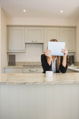 Businesswoman sitting at home behind kitchen island holding tablet computer obscuring face with mug