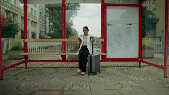 A woman uses a mobile phone when she arrives at a bus stop with a suitcase. She walks along a city street after the rain and talks on her cell phone.