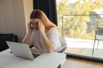 Exhausted woman sitting at desk with laptop, touching her forehead in stress. Concept of burnout, digital fatigue, remote work challenges, mental health, financial worries and technology dependence