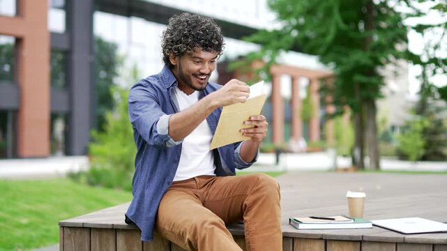 Smiling male student reading letter with great news sitting on a bench in campus near university building. Joyful young handsome man is celebrating success, happy to receive positive good notification