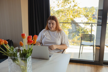 Young woman sitting at desk with laptop in cozy home interior, working remotely or studying online. Concept of freelancing, digital lifestyle, remote work, e-learning, communication, modern technology