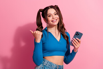 Naklejka premium Cheerful young woman with ponytails holding a smartphone and gesturing positively against a pink background
