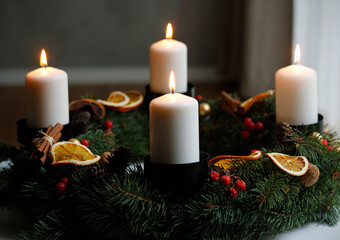 Handmade advent wreath decorated with burning white candles, spruce branches, dried orange slices, cinnamon sticks, pinecones and red berries, traditional Christmas centerpiece glowing warmly