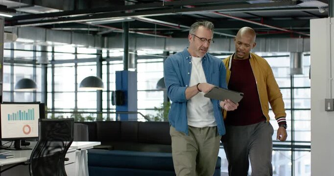 After data request, diverse male colleagues passing office desks tapping tablet reviewing data