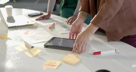Diverse female coworkers placing digital tablet at office desk tapping charts and discussing data - Powered by Adobe
