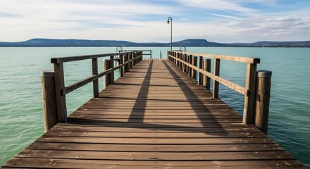 Obraz premium Wooden pier extending into a calm lake with distant mountains under a cloudy sky.