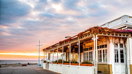 A cozy, romantic cafe on the ocean shore at sunset, illuminated by evening lights. Sea waves, tables, chairs, and lanterns.