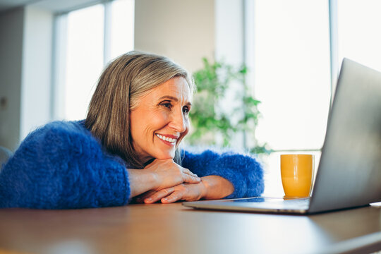 Fototapeta Smiling mature woman in cozy sweater delightfully using a laptop at home while enjoying a relaxed and cheerful moment