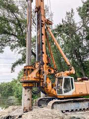 Fototapeta premium Large yellow crane on platformfoundation by water, trees in background, overcast sky, worn appearance, distant figure for scale, daytime, diffused natural light