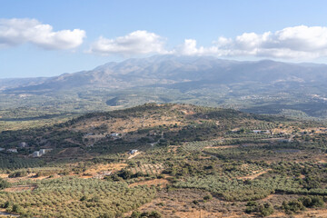 Beautiful panoramic view of the Cretan countryside with mountains, green valleys, olive groves, and a small traditional village under a bright blue sky with scattered clouds. Captured on a sunny day