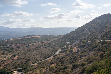 Beautiful panoramic view of the Cretan countryside with mountains, green valleys, olive groves, and a small traditional village under a bright blue sky with scattered clouds. Captured on a sunny day