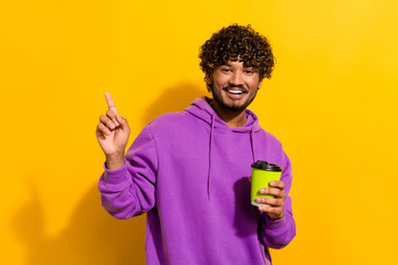 Youthful man in purple hoodie holding coffee pointing upwards on vibrant yellow background with cheerful expression