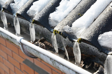 Frozen plastic rain gutter clogged with ice, icicles, and autumn leaves on a house in winter.