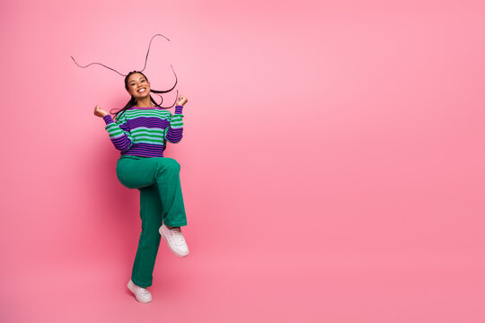 Joyful woman with vibrant striped sweater and green pants leaping enthusiastically on a bright pink background