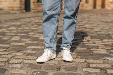 A person is standing on a cobblestone street, wearing jeans and white sneakers during daylight hours