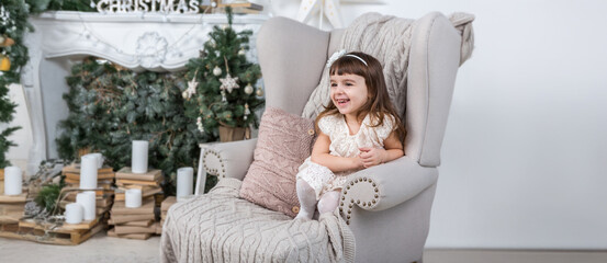 A girl sits in a large armchair in a bright living room decorated for New Year and Christmas. The...