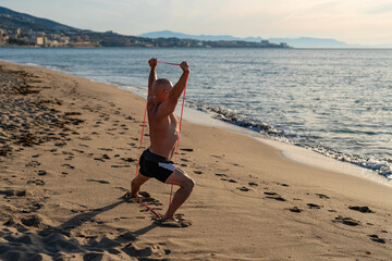 Strong bald man exercising with red resistance bands on sandy beach, raising arms above head in wide stance during outdoor workout near sea waves at sunset