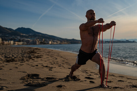 Strong bald man exercising with red resistance bands on sandy beach at sunset, doing forward lunge workout with city skyline and mountains in the background