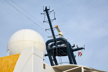 A close-up of the navigation and communication mast of a modern vessel, featuring a large satellite radome, radar scanner, and antennas against a sky.