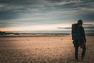 Person walking on a sandy beach with a backpack during twilight hours under a cloudy sky