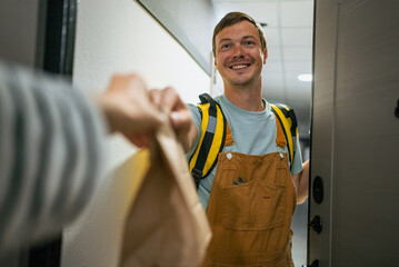 Courier man wearing a backpack giving a brown paper bag containing takeaway food to a person's outstretched hand at their front door, offering convenient home delivery