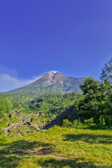 Fototapeta premium A close-up view of Mount Merapi from a distance with trees in the foreground. The panoramic beauty of Mount Merapi on a clear, midday day is clearly visible from a distance.