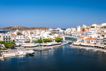 Obraz premium A serene waterfront town in Crete, Greece, featuring whitewashed buildings, terracotta rooftops, a small harbor with boats, and a curved pedestrian bridge.
