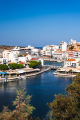 Fototapeta premium A serene coastal town Agios Nikolaos on Crete, Greece, featuring a lagoon, a small bridge, white and pastel buildings, waterfront cafes, and a bright blue sky.