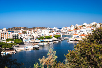 Obraz premium A serene waterfront town Agios Nikolaos in Crete, Greece, featuring whitewashed buildings, red tiled roofs, a small harbor with boats, and a backdrop of rolling hills.